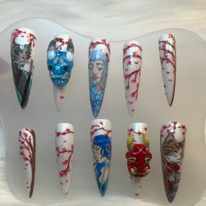 May include: Set of ten long, white, pointy fake nails with different designs. The designs include a cat, a dragon, a woman, cherry blossoms, and a demon mask.