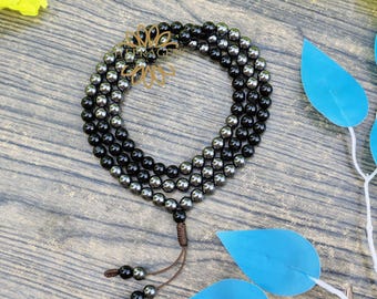 May include: A three-strand beaded necklace with black and silver beads. The necklace is arranged in a circular shape, with a gold-colored floral accent at the top. A brown tassel hangs from the bottom. The beads are approximately 0.3 inches in diameter.