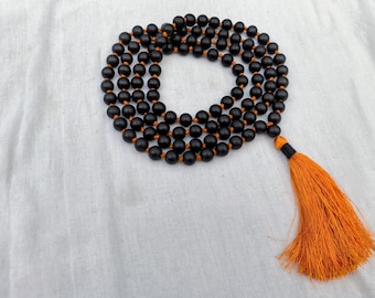 Puede incluir: Un collar de cuentas de madera negra con una borla naranja. Las cuentas están ensartadas en un cordón marrón. La borla es de color naranja brillante con un acento negro. El collar se exhibe sobre una tela blanca.