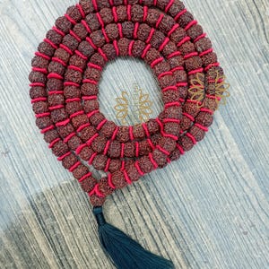 May include: A mala necklace featuring brown beads threaded on red cord, with a dark teal tassel. The beads are arranged in a circular design. The necklace is presented on a light blue wooden surface.