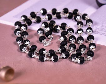 May include: A necklace featuring black beads and silver-coloured detailing. The beads are spherical and regularly spaced, connected by a fine chain. The necklace is presented on a pink surface, with a book visible in the background.
