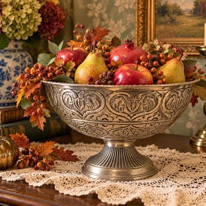 May include: A silver-toned metal pedestal bowl featuring an embossed leaf pattern around the exterior. The bowl is filled with a collection of red apples, yellow-green pears, a pomegranate, and various autumn-colored berries and foliage.
