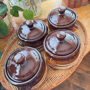 May include: A set of four round ceramic crocks with glossy dark brown lids and bodies, featuring a lighter tan band around the base. Each crock has a small side handle and a knob on the lid, all arranged together on an oval woven rattan tray.