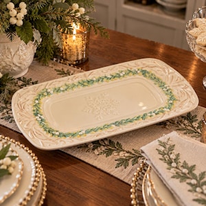 May include: A rectangular cream-colored ceramic serving platter featuring an embossed snowflake design in the center and a decorative border of green leaves, yellow accents, and white berries along the rim with a scalloped edge.