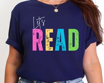 May include: Navy blue t-shirt with the words "Let's READ" in colorful, distressed lettering. The text is in pink, yellow, teal, and green. The shirt has short sleeves and a crew neck.