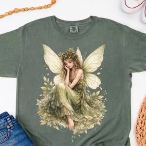 May include: Moss green t-shirt featuring a detailed illustration of a fairy with large wings, a floral crown, and a flowing green dress. The fairy is seated amidst leaves and flowers. The shirt is paired with denim shorts and white sneakers.