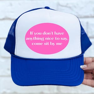 May include: A blue and white trucker hat with a pink oval that reads, "If you don't have anything nice to say, come sit by me." The hat has a blue brim and mesh backing.