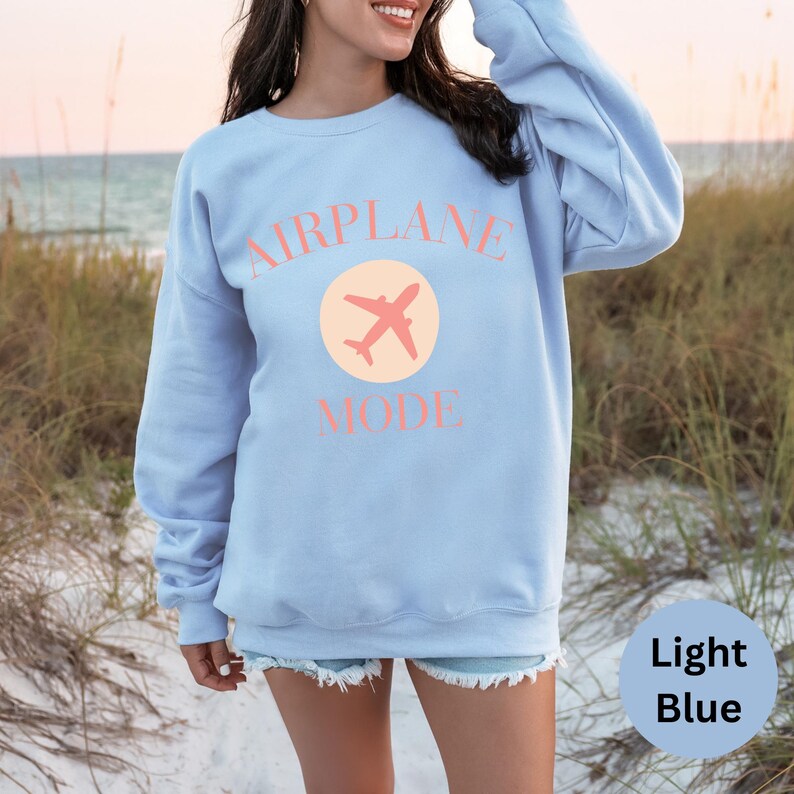 Light blue colored crewneck sweatshirt. Pink colored letters in large all caps that read AIRPLANE MODE. Graphic printed in middle of the shirt is of a an airplane; similar to the airplane mode symbol used on a cellular phone. Model is wearing an oversized fit. Various colors and sizes available. Perfect cozy sweatshirt for anyone in vacay mode, or gift for a traveler.
