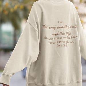 Puede incluir: Sudadera color crema con el texto "Yo soy el camino, la verdad y la vida. Nadie viene al Padre, sino por mí. Juan 14:6" impreso en la espalda en fuente marrón. La sudadera tiene mangas largas y cuello redondo.