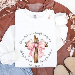 May include: White sweatshirt with a watercolour illustration of a wooden cross and a pink bow. The design includes the text "She is clothed with strength and dignity" and "She laughs without fear of the future" with "Proverbs 31:25" below.