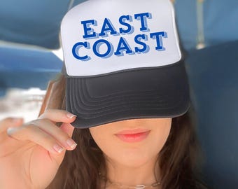 Op de afbeelding: Een zwart-witte truckerpet met de woorden "EAST COAST" in blauwe blokletters. De pet wordt door een persoon gedragen. De pet heeft een wit voorpaneel en een zwarte klep en mesh rug.