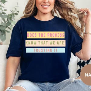 Könnte beinhalten: Marineblaues T-Shirt mit dem Text "DOES THE PROCESS KNOW THAT WE ARE TRUSTING IT" in farbigen rechteckigen Blöcken. Das Shirt hat eine marineblaue Farbe und einen lässigen Stil.