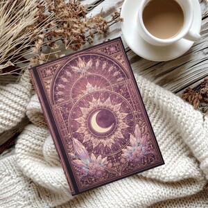 Peut inclure: Un journal violet avec un motif céleste représentant un croissant de lune, un soleil et des accents de cristal. Le journal est ouvert sur une couverture en tricot blanc, avec une tasse de café et des plantes séchées en arrière-plan.