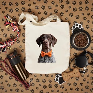 May include: A cream-colored tote bag featuring a German Shorthaired Pointer wearing an orange bow tie. The dog is brown and white with a white stripe on its head. The background is brown with black paw prints. Other dog accessories are also visible.