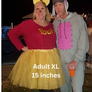 Pu&ograve; includere: Una donna indossa una maglietta rossa con la scritta "Pooh" e una gonna di tulle dorata. Indossa una fascia per capelli con orecchie da topo. La gonna &egrave; lunga 38 cm ed &egrave; di taglia XL per adulti. La donna &egrave; in piedi accanto a un uomo che indossa una felpa grigia con una toppa rosa sul davanti. L'uomo indossa una fascia per capelli con orecchie da coniglio.