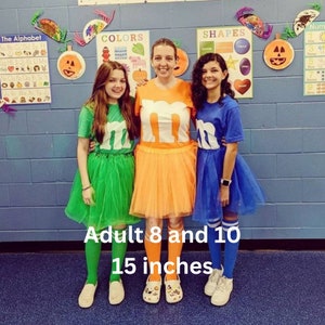 Pu&ograve; includere: Tre persone vestite con costumi della lettera M con una gonna a tut&ugrave;. Le gonne sono verde, arancione e blu. Il testo "Adult 8 and 10 15 inches" &egrave; scritto sotto le persone.