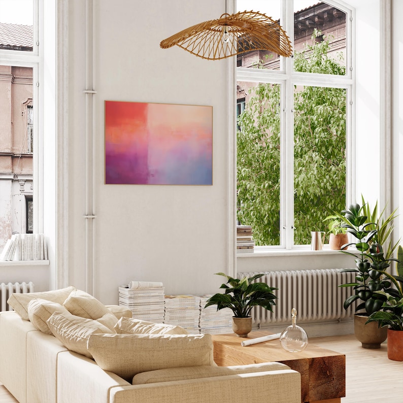 May include: A living room interior with a white couch, a wooden coffee table, and a large abstract painting on the wall. The painting features a gradient of pink, orange, and blue colors. There are plants in pots and a wicker pendant light hanging from the ceiling.