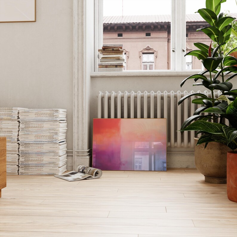 May include: A framed abstract painting with a gradient of pink, orange, and purple hues. The painting is hanging on a white wall in a room with a wooden floor and a large window.