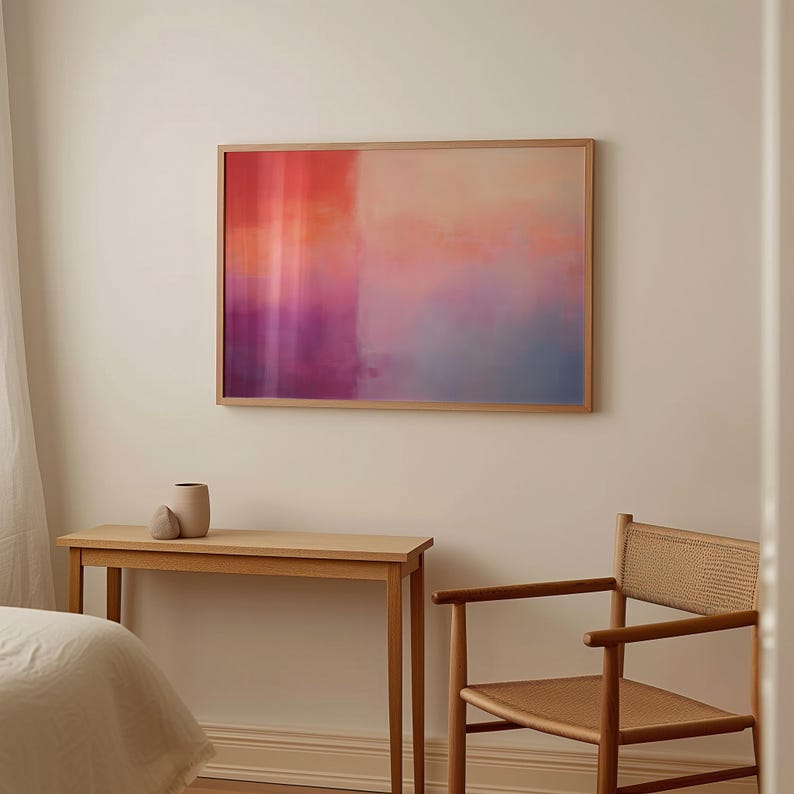May include: A framed abstract painting with a gradient of pink, orange, and blue colors. The painting is hanging on a white wall above a wooden table and chair.
