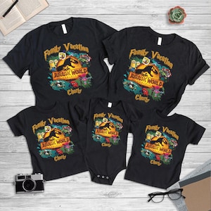 Puede incluir: Una colección de camisetas negras y un body para bebé con el texto "Family Vacation" y "Jurassic World" con un diseño de dinosaurio. Las camisetas son de varios tamaños, adecuadas para una familia. También se ve una cámara y gafas.