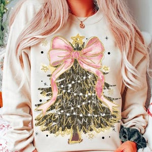 Può includere: Una felpa color crema con un design a forma di albero di Natale. L'albero è adornato con un fiocco rosa, dettagli dorati e luci a catena. Il design include stelle e fiocchi di neve dorati, creando un tema festivo natalizio.