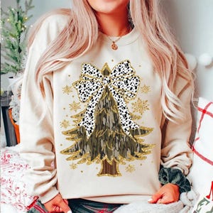 Può includere: Una felpa beige con un design ad albero di Natale. L'albero è decorato con un motivo mimetico e un fiocco a pois bianchi e neri. Accenti di glitter dorati e fiocchi di neve aggiungono brillantezza all'abbigliamento festivo.