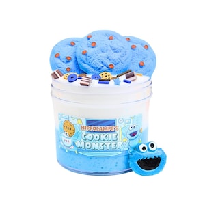 Pode incluir: Um recipiente de plástico transparente preenchido com slime azul e branco, coberto com três decorações azuis em forma de biscoito com granulado laranja. O recipiente tem a inscrição "Hippocampe's Cookie Monster" e uma pequena figura do Monstro das Bolachas.