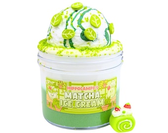 May include: A clear plastic jar filled with green and white slime, topped with a white, textured substance, green sprinkles, and leaf-shaped decorations. The jar has a label that reads "Hippocampe's Matcha Ice Cream." A small, green, roll cake with white frosting and red strawberry decorations sits beside the jar.