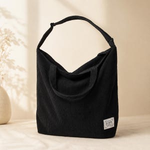 May include: A black corduroy tote bag with a single shoulder strap and a smaller handle. The bag has a rectangular shape and a small, rectangular label on the lower right corner.
