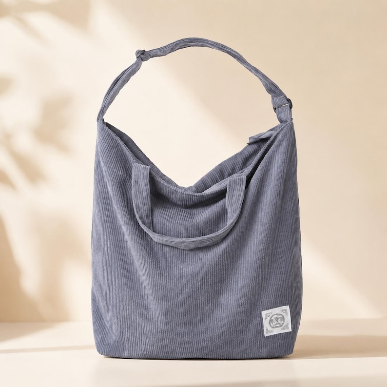 May include: A dusty blue corduroy tote bag with a single shoulder strap and a smaller handle. The bag has a rectangular shape with a small label on the front. The corduroy fabric has vertical ridges.