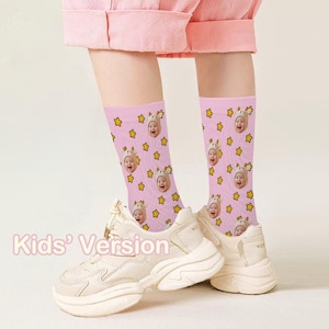 May include: Pink socks with a repeating pattern of a baby's face wearing a hat and yellow stars. The socks are worn with white sneakers. The text "Kids' Version" is visible.