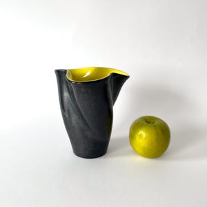 May include: A black ceramic vase with a yellow interior and a twisted design. A green apple sits to the right of the vase. The vase has a unique, sculptural shape and a smooth finish.