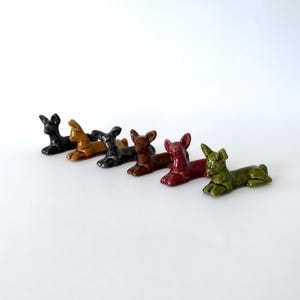 May include: A collection of six ceramic dog figurines in various colors, including black, gold, dark gray, brown, burgundy, and green. The dogs are in a reclining pose, with their front legs extended. The figurines are likely decorative items.