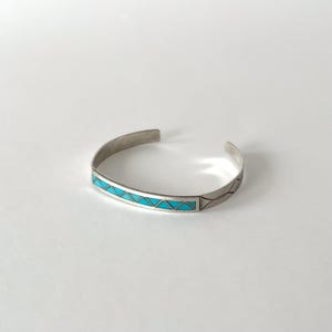 Peut inclure: Un bracelet manchette en argent doté d'une incrustation horizontale centrale composée de segments triangulaires turquoise vif. La bande métallique présente une finition lisse et polie avec de fines lignes gravées sur les côtés.