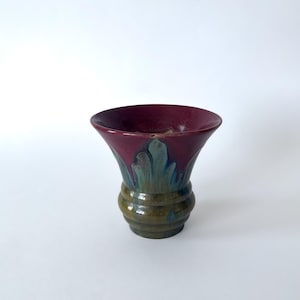 May include: A ceramic vase featuring a wide, flared rim with a deep burgundy glaze that transitions into a drip pattern of teal and blue, ending in a rounded, ribbed base with earthy olive green and brown tones throughout the lower section.
