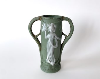 Könnte beinhalten: Grüne Keramikvase mit zwei Henkeln und einem weißen Relief einer Frau, die einen Vogel hält. Die Vase hat eine matte Oberfläche und ein detailliertes Blumendesign um den Sockel und die Figur der Frau.