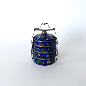 Pode incluir: Um recipiente cilíndrico de metal para alimentos de quatro níveis com um acabamento em esmalte azul vibrante e padrões florais multicoloridos complexos. O recipiente inclui uma alça de metal em tom de latão e um botão central na tampa.