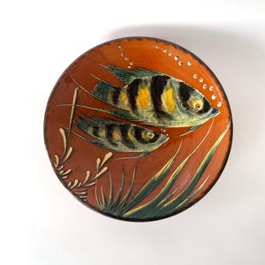 May include: Round ceramic plate with a hand-painted design featuring two fish in a vibrant orange and green underwater scene. The fish have black and yellow stripes. Decorative bubbles and aquatic plants complete the design.