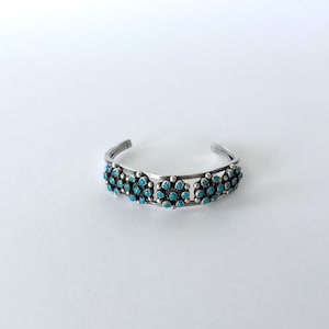 Peut inclure: Un bracelet manchette en argent orné de cinq grappes circulaires de petites pierres turquoise bleu vif. La bande métallique est divisée en deux fils parallèles, et chaque grappe de turquoise est entourée de minuscules perles d'argent.