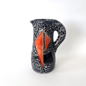 Peut inclure: Un pichet en céramique avec une finition émaillée mouchetée noire et blanche texturée. Le corps est décoré de deux formes triangulaires géométriques orange proéminentes et d'un accent triangulaire noir sur le bas avec une anse.