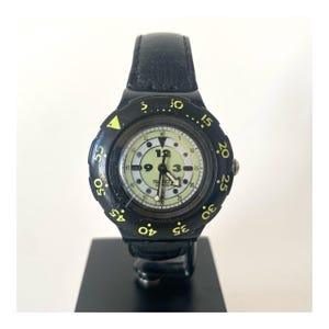 以下が含まれることがあります： 黒の革バンドと黄黒のベゼルが付いた、黒いスウォッチの腕時計。文字盤は白で、黒の数字と針が付いています。時計には3時位置に日付表示窓があります。