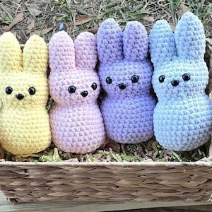 May include: Four crocheted bunny plushes in a wicker basket. The bunnies are yellow, lavender, light purple, and blue.