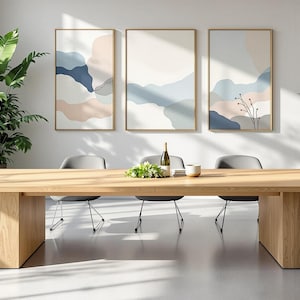 Può includere: Un lungo tavolo da pranzo in legno con sei sedie in una sala da pranzo moderna. Il tavolo ha una finitura in legno chiaro e le sedie sono grigie. Ci sono tre stampe d'arte astratta sul muro dietro il tavolo. Le stampe sono in tonalità di blu, rosa e bianco.