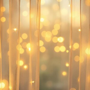Puede incluir: Una cortina blanca transparente con luces de hadas blancas cálidas enhebradas a través de ella. Las luces crean un efecto suave y brillante contra la cortina. El fondo está desenfocado, lo que sugiere una ventana con la luz del sol brillando a través.