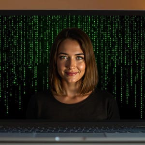以下が含まれることがあります： 女性がラップトップの画面を通してカメラを見ている。画面には緑色のマトリックスのようなパターンが表示されている。