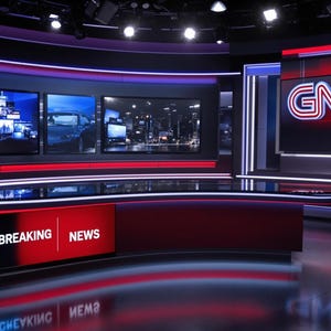 May include: A television studio news set with red and black accents. Three large screens display breaking news footage. The set features a curved desk with a "BREAKING NEWS" sign. The logo "GN" is displayed on a screen in the background.