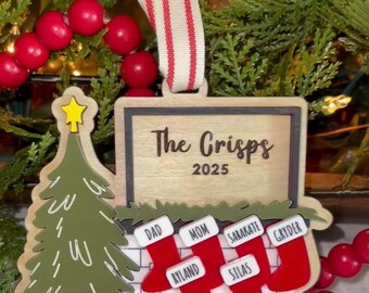 May include: A wooden Christmas ornament featuring a Christmas tree and a fireplace with red stockings. The ornament reads "The Crisps 2025" with names in the stockings. It is decorated with a red and white striped ribbon.