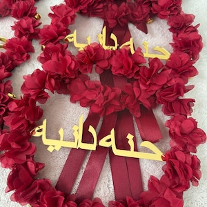 May include: Red floral wreath with gold lettering and burgundy ribbons. The wreath is made of layered fabric flowers and small gold bells. The gold lettering appears to be Arabic script.