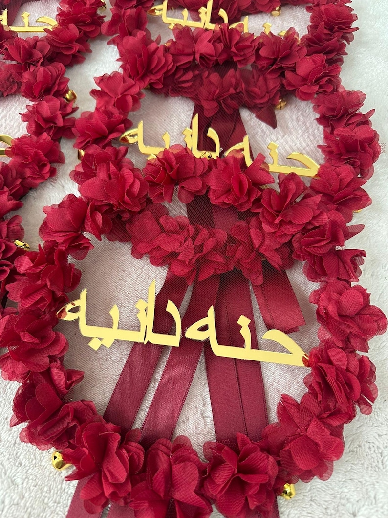 May include: Red floral wreath with gold lettering and burgundy ribbons. The wreath is made of layered fabric flowers and small gold bells. The gold lettering appears to be Arabic script.