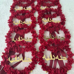 May include: A collection of burgundy floral wreaths with gold lettering. Each wreath is adorned with small, fabric flowers and long, burgundy ribbons. The gold lettering appears to be Arabic script. The wreaths are likely for a special occasion.